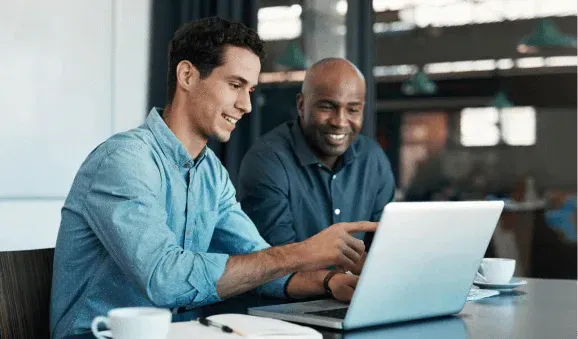 Two men sitting at a table, engaged in discussion while using a laptop.