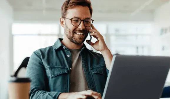 A smiling man with glasses talks on the phone while using a laptop.